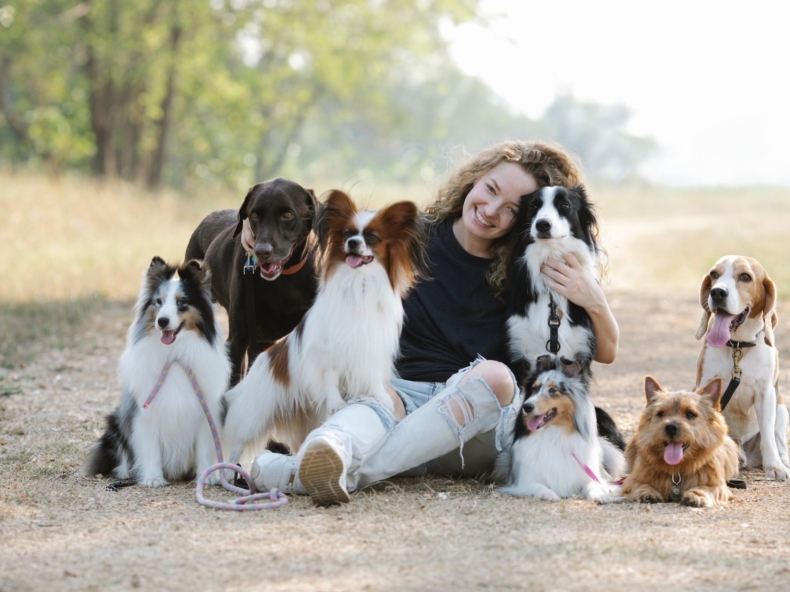 Een jonge vrouw die glimlacht naar de camera en omringt is door verschillende honden